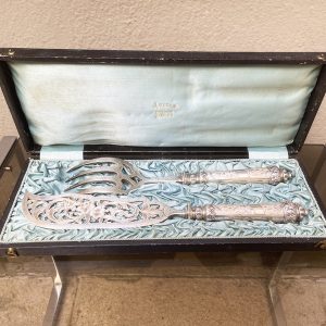 Un ensemble raffiné qui rappelle les grands dîners d'antan : cette fourchette et ce couteau de service à poisson vintage en argent ( ?) reposent encore dans leur écrin d'origine, gainé de cuir noir et doublé de satin bleu pâle estampillé « Julien Brest ». Les manches, de section fuselée, sont ornés d'un fin décor gravé de feuillages et de rinceaux typique du style Louis XVI, rehaussé de frises de feuilles d'acanthe et de perles aux extrémités. La lame du couteau se distingue par un magnifique travail ajouré, dévoilant un bouquet de fleurs entouré de volutes végétales — un ouvrage d'orfèvrerie d'une grande délicatesse. La fourchette, aux dents largement échancrées en cœur, répond à ce même vocabulaire décoratif avec une gravure soignée sur le col. Quelques signes d’usure normale. (boite) LO 34,5 x LA 13,5 x H 4,5 cm (couverts) LO 27,5 à 31 cm