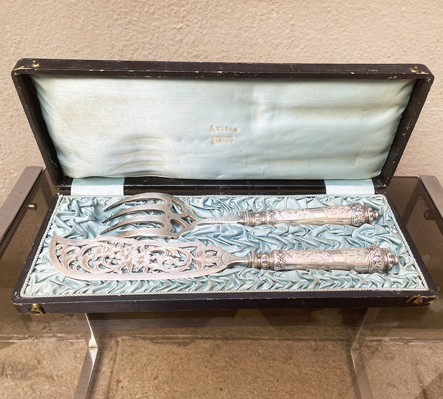 Un ensemble raffiné qui rappelle les grands dîners d'antan : cette fourchette et ce couteau de service à poisson vintage en argent ( ?) reposent encore dans leur écrin d'origine, gainé de cuir noir et doublé de satin bleu pâle estampillé « Julien Brest ». Les manches, de section fuselée, sont ornés d'un fin décor gravé de feuillages et de rinceaux typique du style Louis XVI, rehaussé de frises de feuilles d'acanthe et de perles aux extrémités. La lame du couteau se distingue par un magnifique travail ajouré, dévoilant un bouquet de fleurs entouré de volutes végétales — un ouvrage d'orfèvrerie d'une grande délicatesse. La fourchette, aux dents largement échancrées en cœur, répond à ce même vocabulaire décoratif avec une gravure soignée sur le col. Quelques signes d’usure normale. (boite) LO 34,5 x LA 13,5 x H 4,5 cm (couverts) LO 27,5 à 31 cm